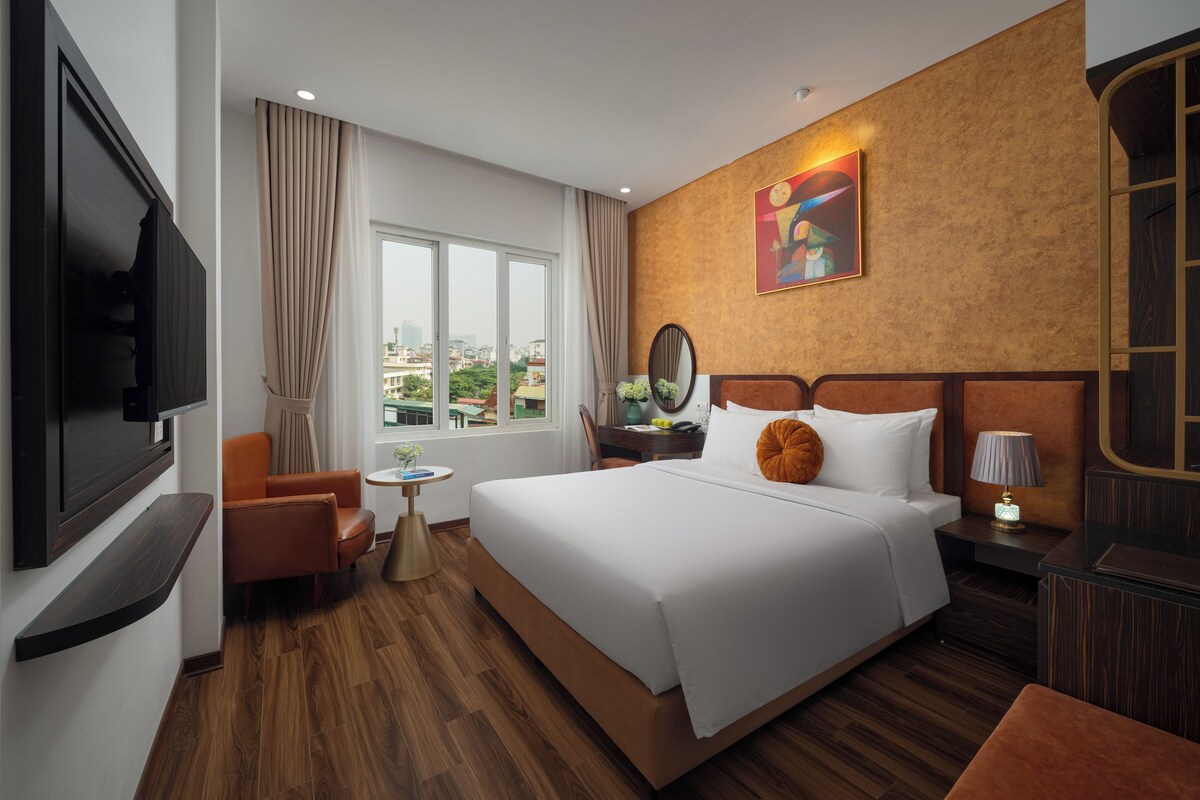 Ein Hotelzimmer mit einem großen Bett, einem Fernseher, einem Schreibtisch mit Lampe und Blick auf die Stadt durch das Fenster.