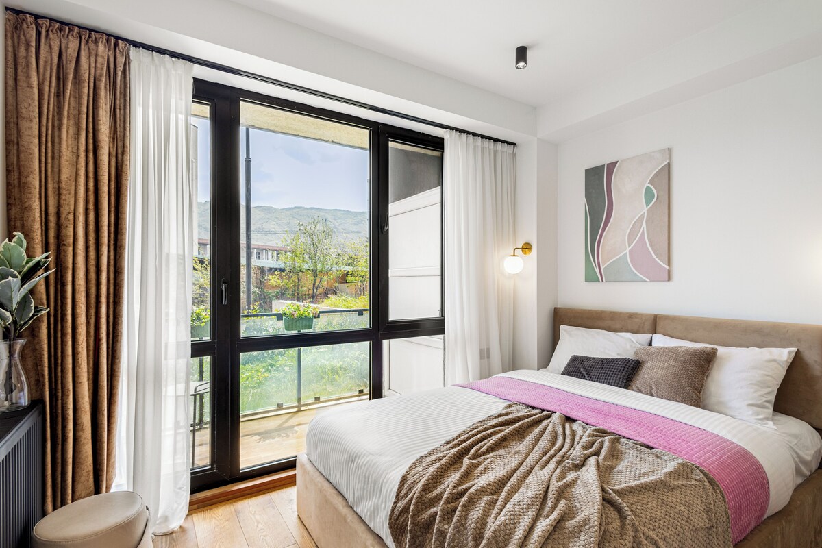 A bedroom with a bed, a window with curtains, a bedside table, and a wall-mounted light.