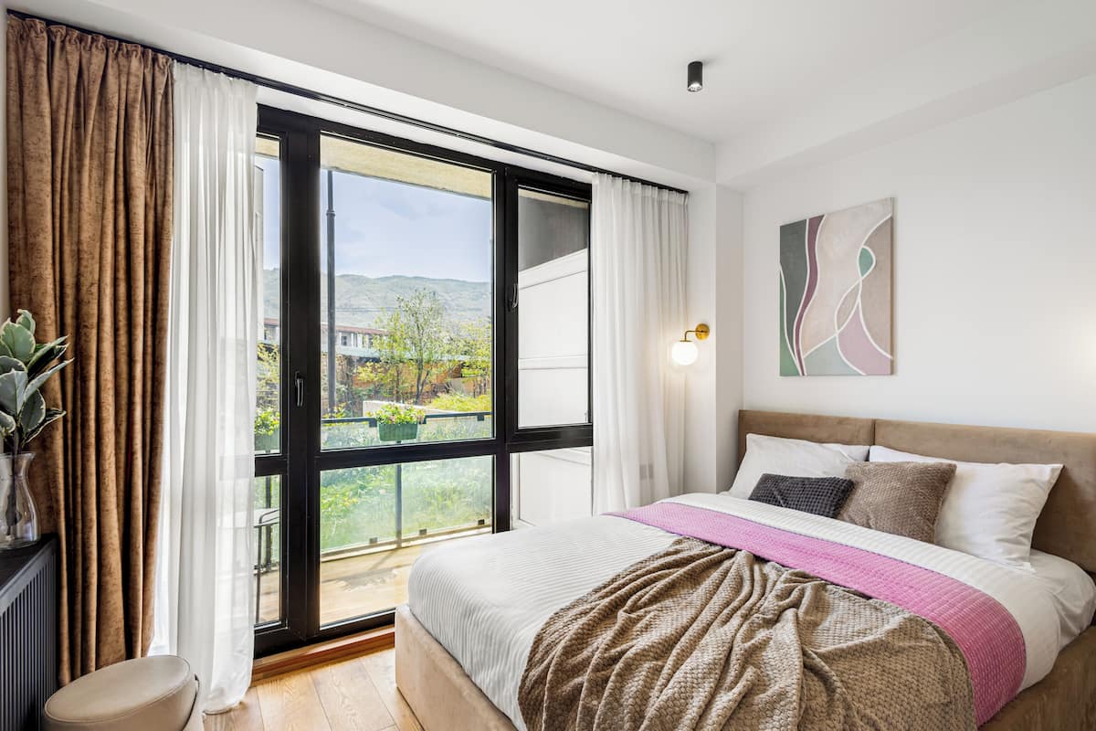 Ein Schlafzimmer mit Bett, Vorhängen an einem Fenster, einem Nachttisch und einer Wandleuchte.