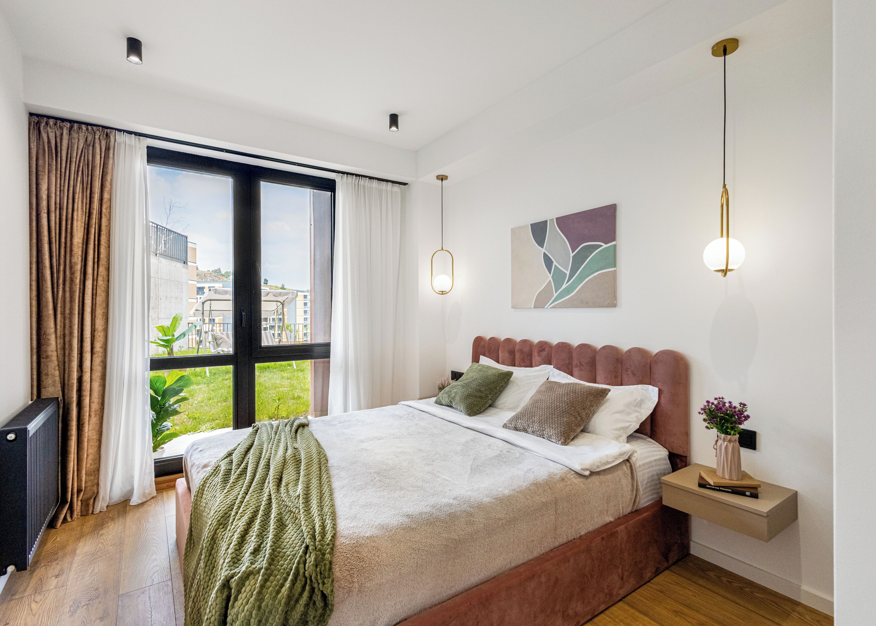 Ein modernes Schlafzimmer mit großem Fenster, einem Bett mit vielen Kissen, einem Nachttisch mit Lampe und einem Wandbild.