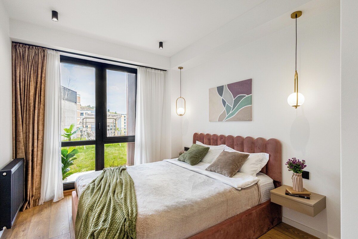 A modern bedroom with a large window, a bed with multiple pillows, a bedside table with a lamp, and a wall-mounted artwork.
