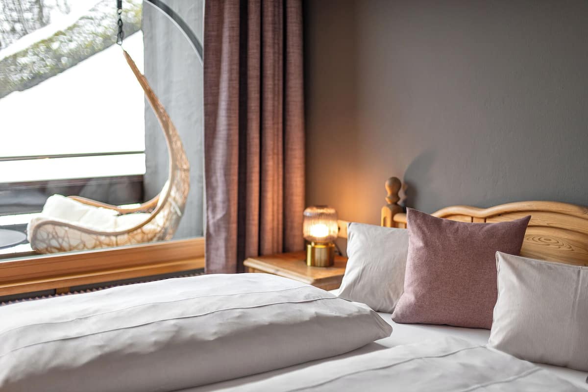 A neatly made bed with pillows, a bedside lamp, a wooden headboard, and a window with curtains.