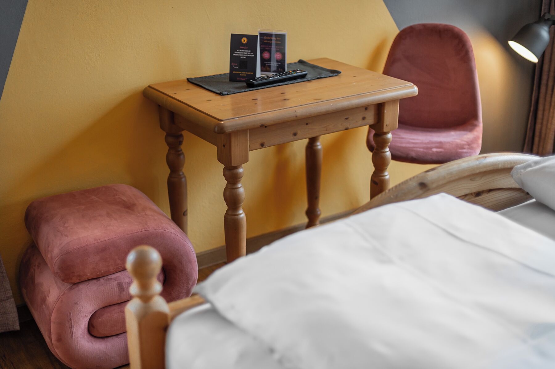 Una mesita de noche de madera con un control remoto y folletos informativos, una silla rosa, un cabecero de madera y una lámpara.