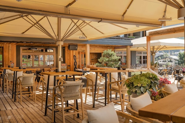 An outdoor cafe with wooden tables and chairs, umbrellas, and potted plants.