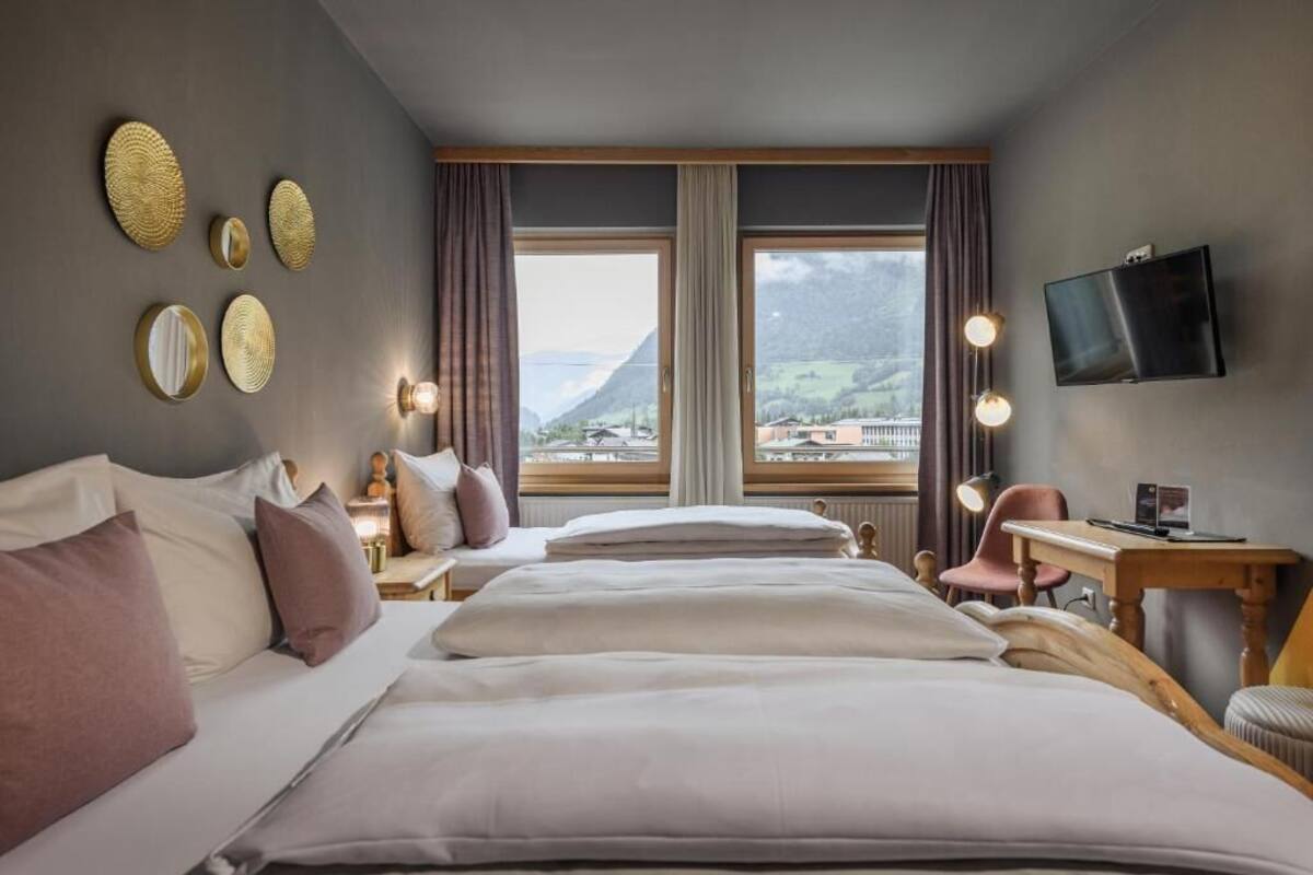 A hotel room with two beds, a desk, a chair, a TV, and a large window with a mountain view.