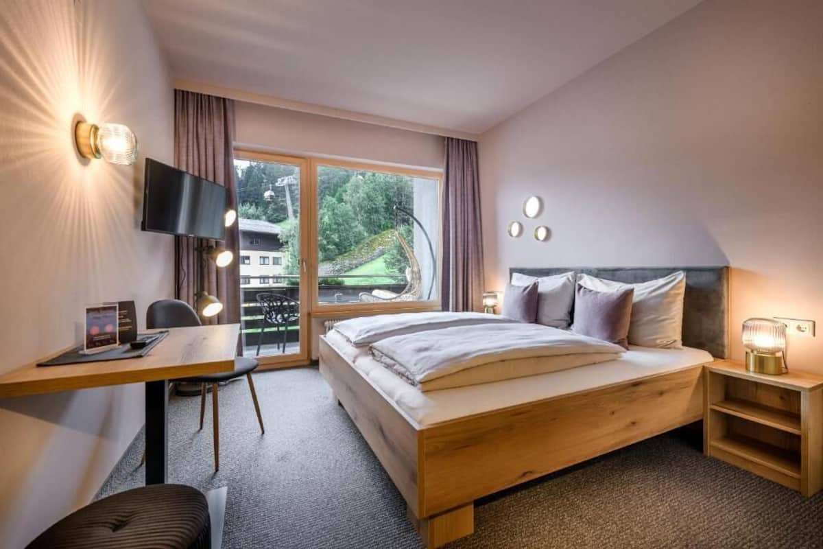 A hotel room with a bed, a desk, a TV, and a window with a view of greenery.