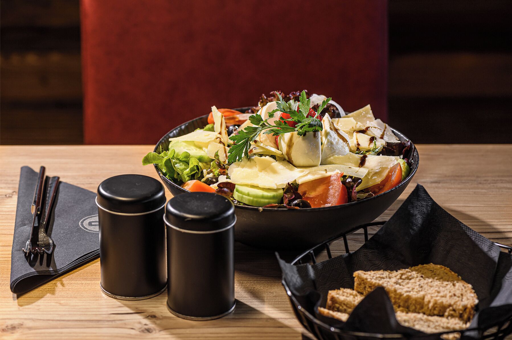 A wooden table with a salad bowl, two salt shakers, a napkin, and a basket of bread.