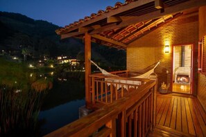 Una terraza de madera con una hamaca, un edificio de ladrillo con chimenea y vista a un lago al anochecer.