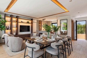 A modern dining area with a long table set for six, surrounded by comfortable chairs, a TV mounted on the wall, and a large window offering a view of the outdoors. - Luxury Golf Townhouse in Aloha with Pool & Garden (Marbella)
