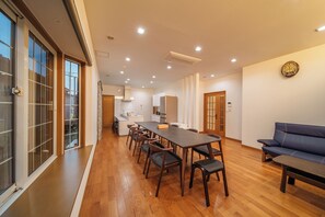 A spacious living area with a long dining table, multiple chairs, a sofa, and a kitchen in the background. - KINNOKA YUNOYADO (Yufu)