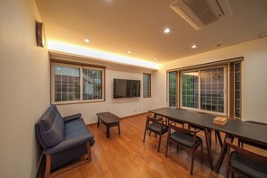 A room with a sofa, a dining table with chairs, a TV mounted on the wall, and large windows. - KINNOKA YUNOYADO (Yufu)