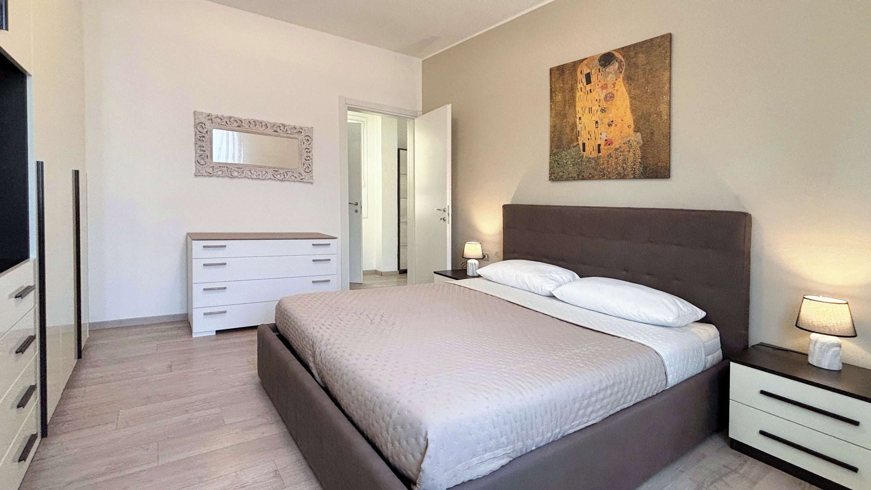 A bedroom with a bed, bedside tables, a dresser, a mirror, and a painting on the wall.