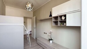 Un pasillo con pared blanca, suelo de madera, una silla blanca, un armario blanco con estantes y una lámpara colgante, además de una puerta que da a otra habitación.
