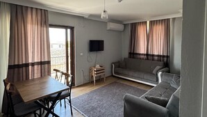 Un salon comprenant un canapé, une table à manger avec des chaises, un téléviseur fixé au mur et une fenêtre avec des rideaux.