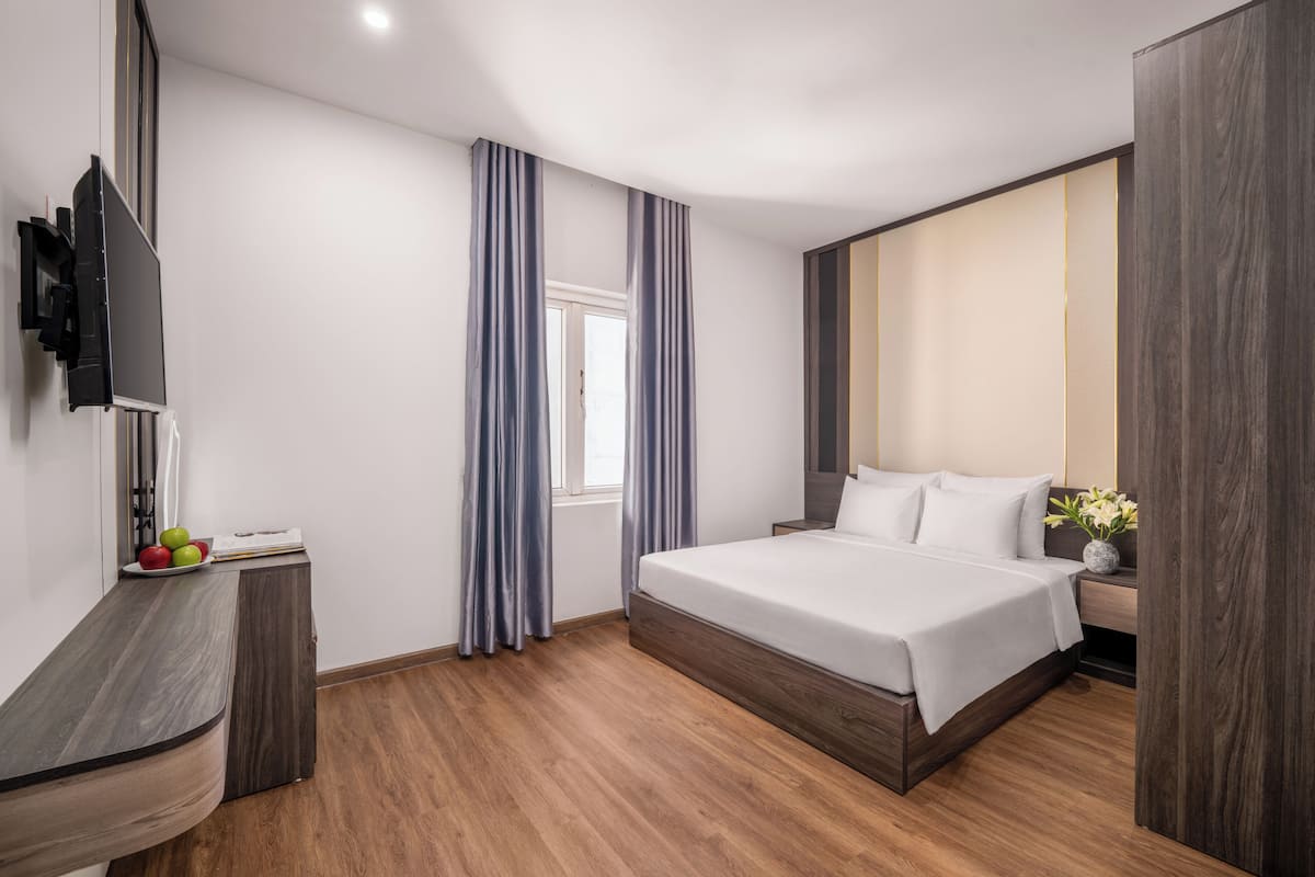 A modern hotel room with a wooden floor, a bed with white linens, a TV mounted on the wall, and a window with curtains.