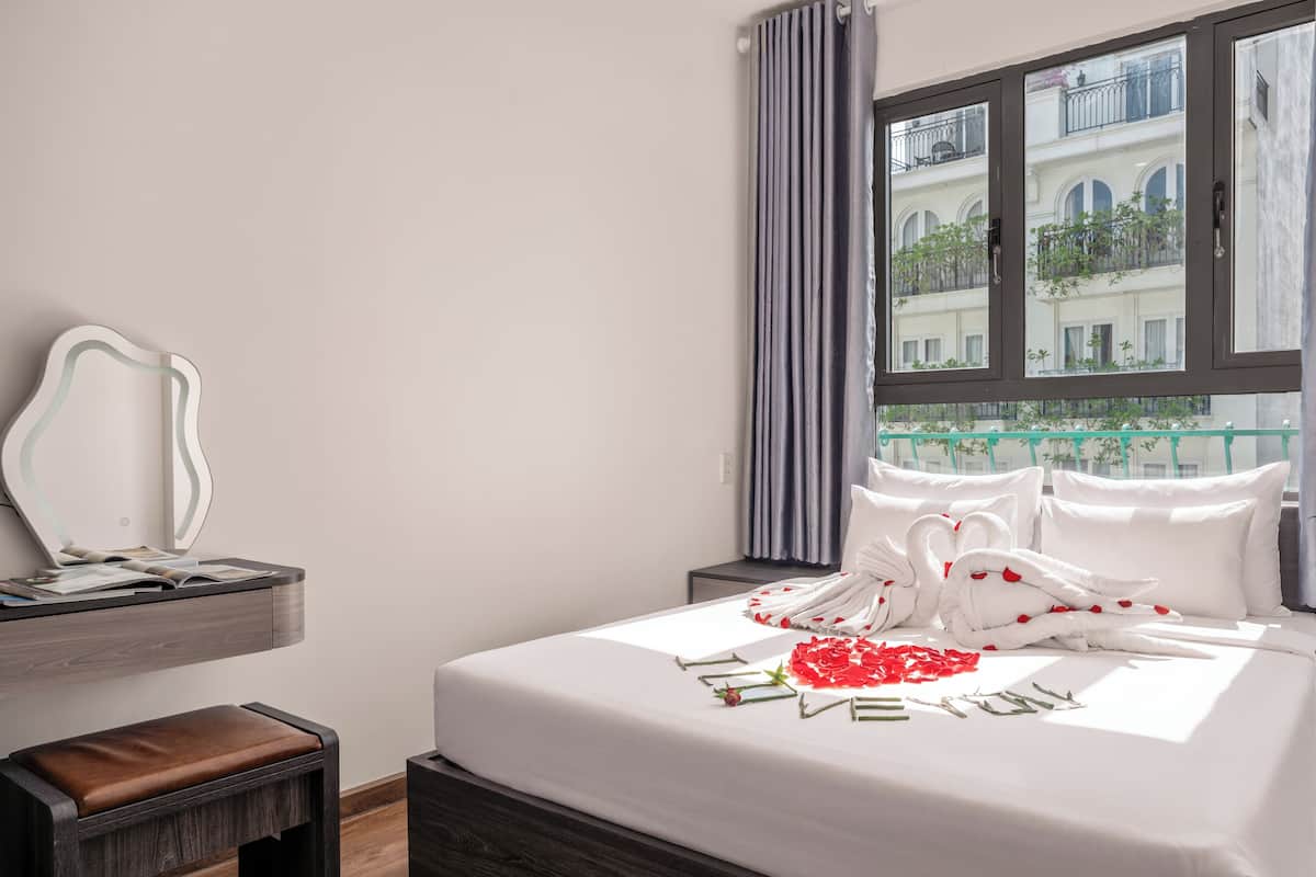 A hotel room with a bed, a nightstand, a bench, a mirror, and a window with a view of buildings.