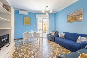 Una sala de estar con un sofá azul, un televisor, una zona de comedor con una mesa y sillas, y un suelo con estampado.