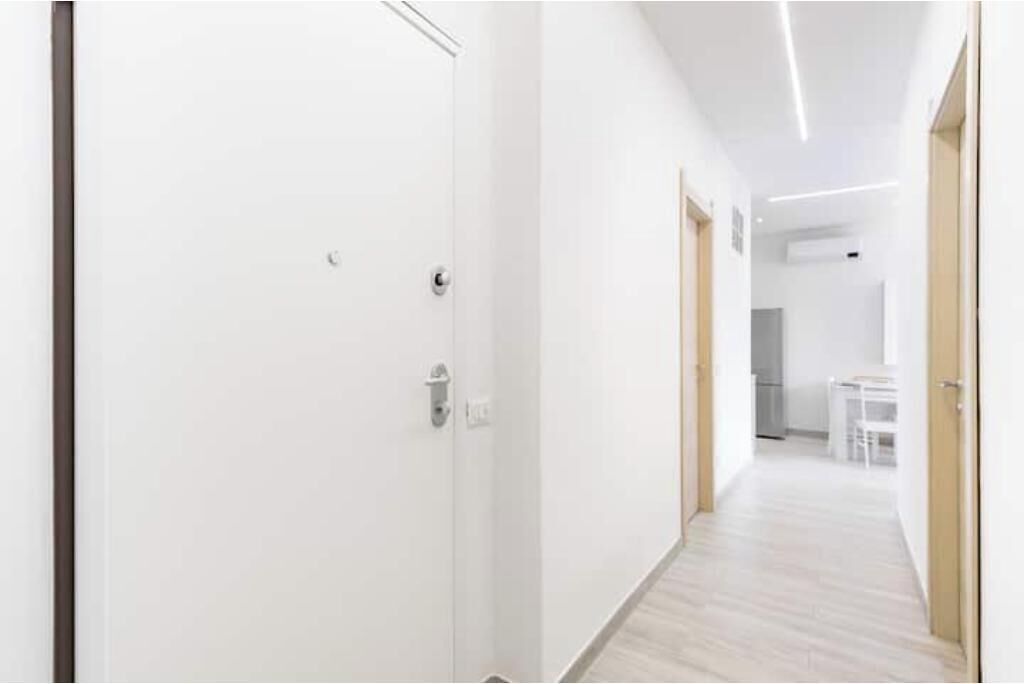 A hallway with white walls and a wooden floor, leading to a kitchen area with white furniture.