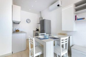 Een kleine keuken met witte kastjes, een magnetron en een fornuis. Er is een eettafel met stoelen en een klok aan de muur.