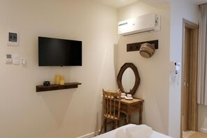 Un dormitorio con televisión de pantalla plana, aire acondicionado, una silla de madera, una mesita con tazas y un espejo.