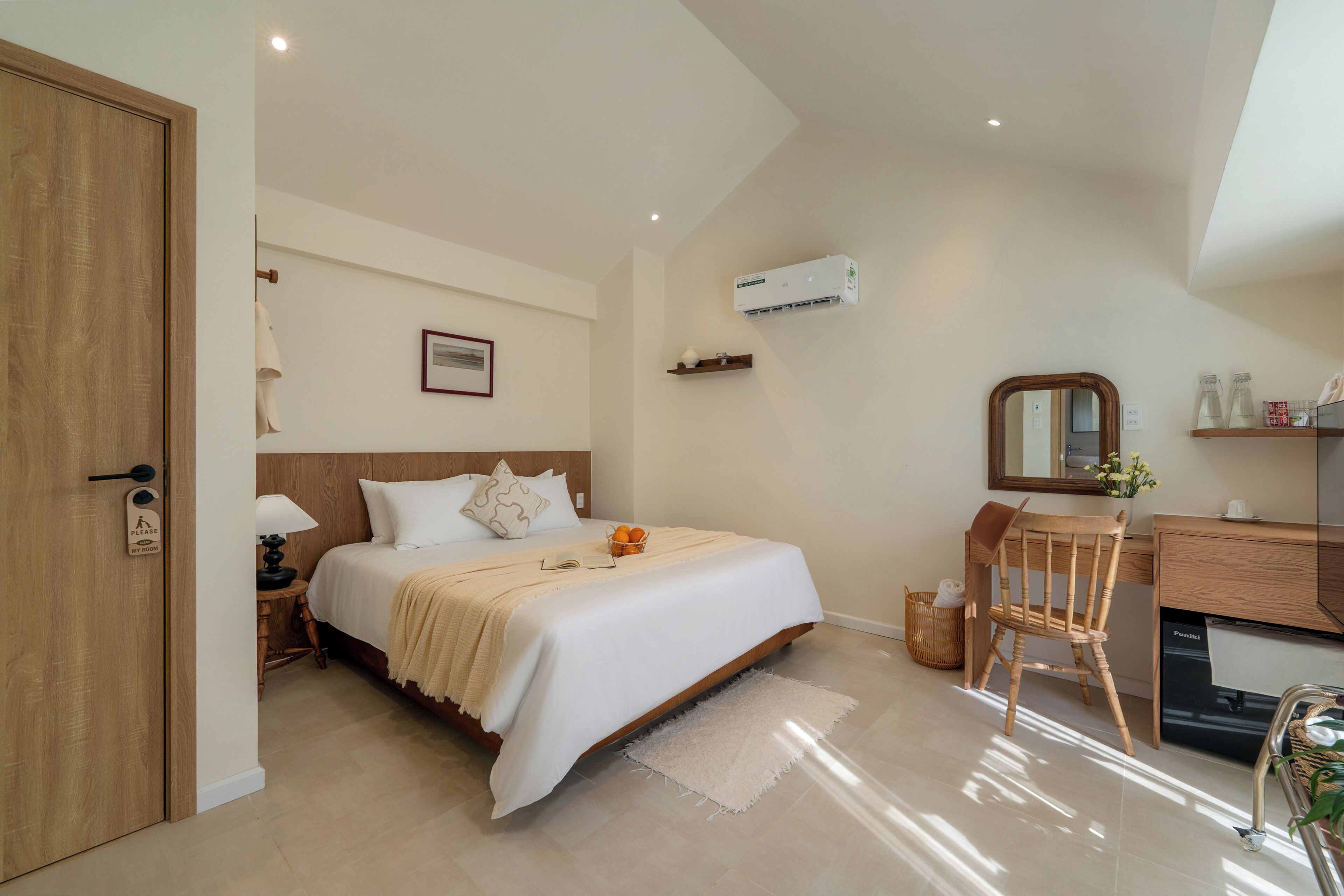 A bedroom with a bed, bedside table, lamp, chair, desk, mirror, and air conditioning unit.