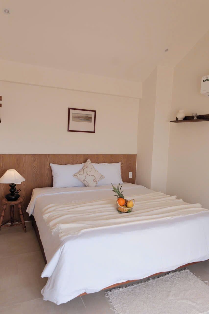 Una cama bien hecha con una colcha blanca, una mesita de noche con una lámpara y una cesta de frutas sobre la cama.