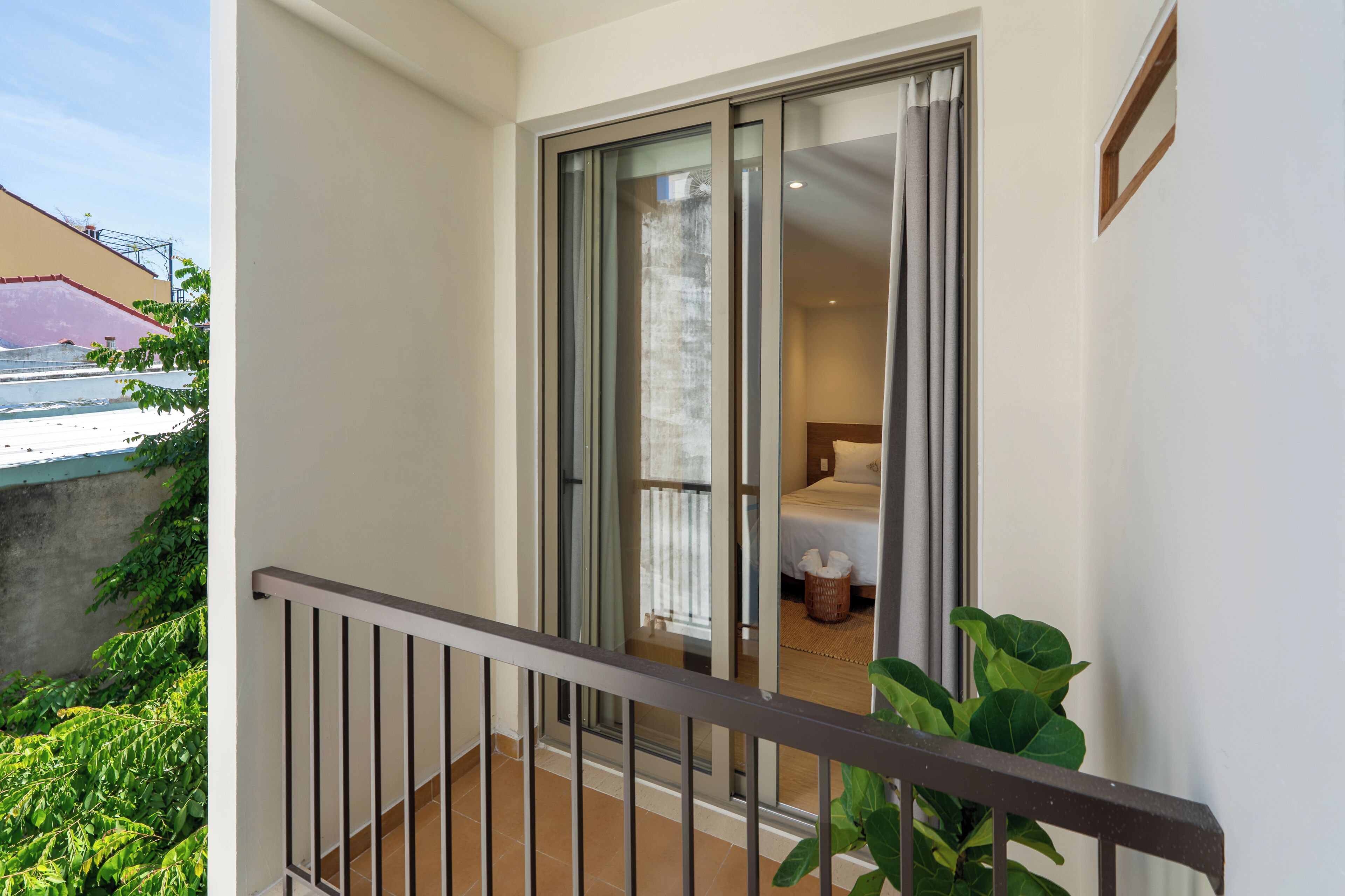 A balcony with a metal railing, a sliding glass door leading to a bedroom with a bed and a nightstand, and a small plant on the right.