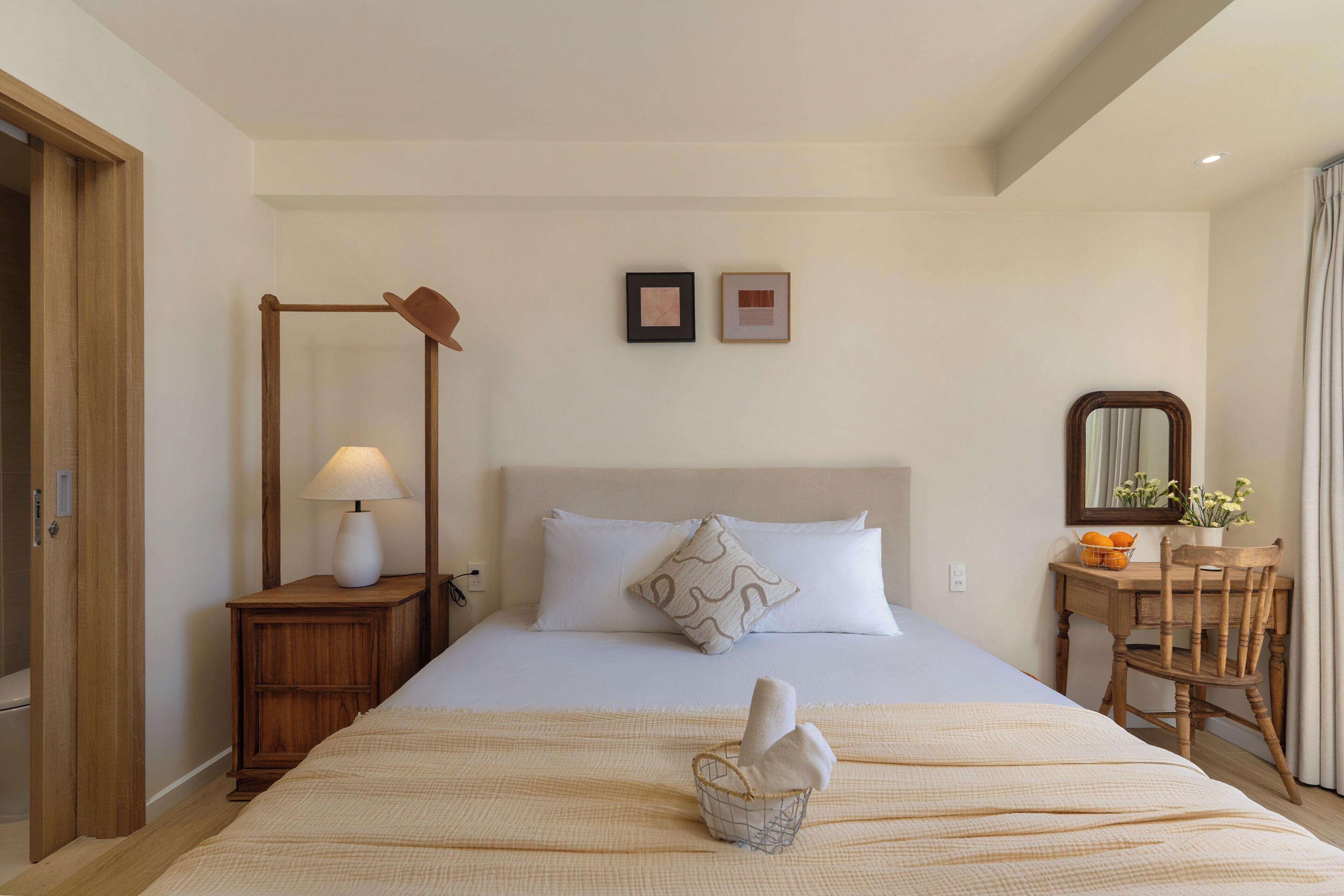 A hotel room with a bed, a nightstand, a lamp, a wooden chair, and a small table with a mirror and flowers.