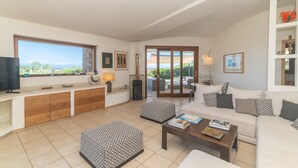 Un salón amplio con sofá, reposapiés, mesa de centro, televisor y vistas a través de amplias ventanas.