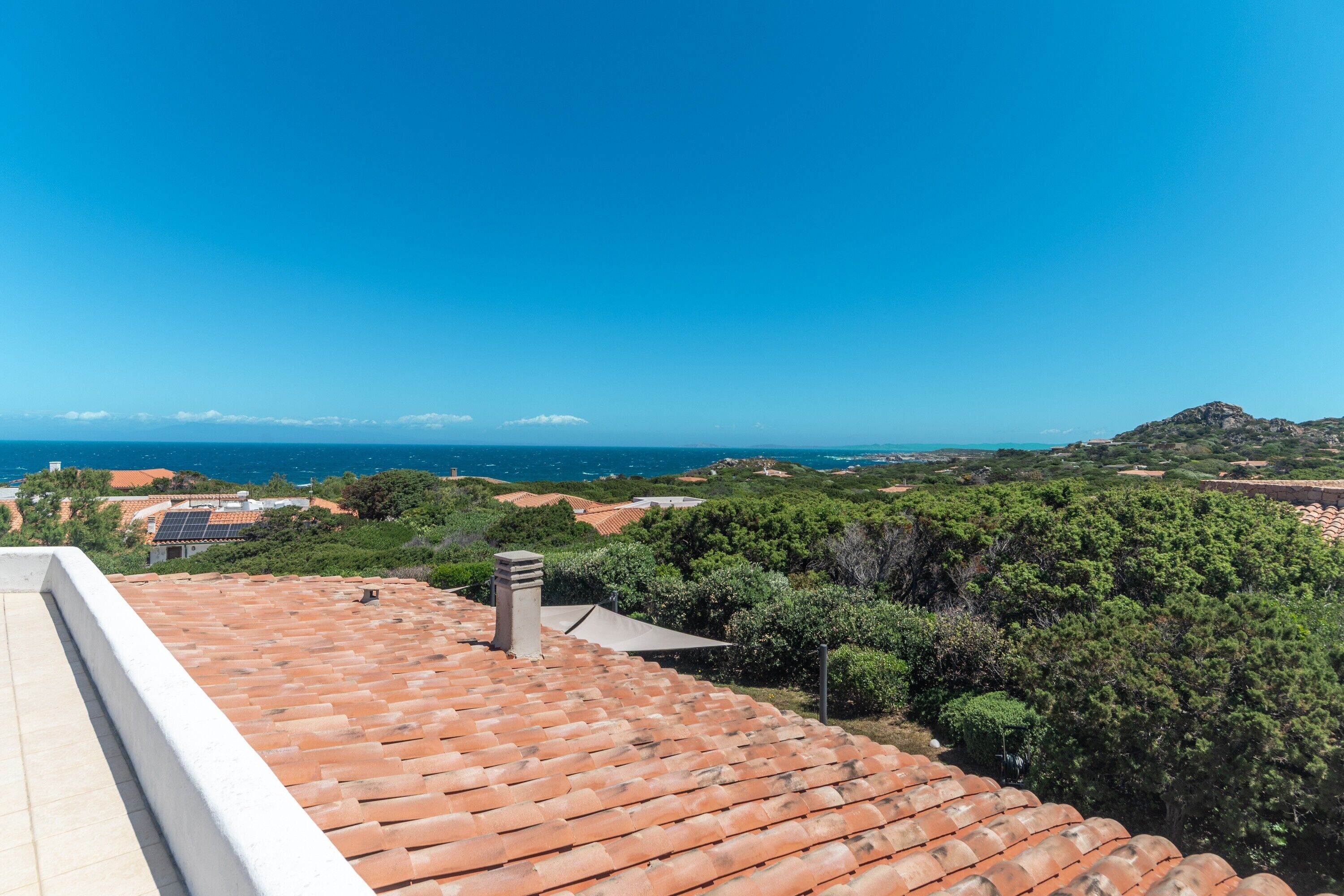 A rooftop terrace with terracotta tiles, a chimney, and a view of the sea and greenery.