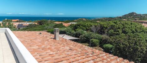 A rooftop terrace with terracotta tiles, a chimney, and a view of the sea and greenery.