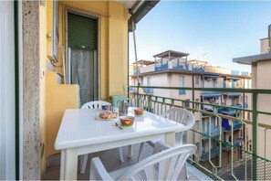 Un balcón con una mesa y sillas blancas, una puerta verde y vistas a edificios cercanos.