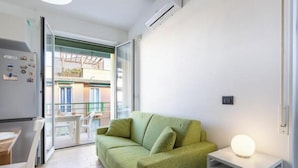 Un salon moderne avec un canapé vert, une petite table avec une lampe et une porte-fenêtre coulissante donnant sur un balcon extérieur.