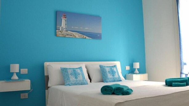 A bedroom with a bed, pillows, a painting of a lighthouse, and a window. - Palau Noal (Alghero)