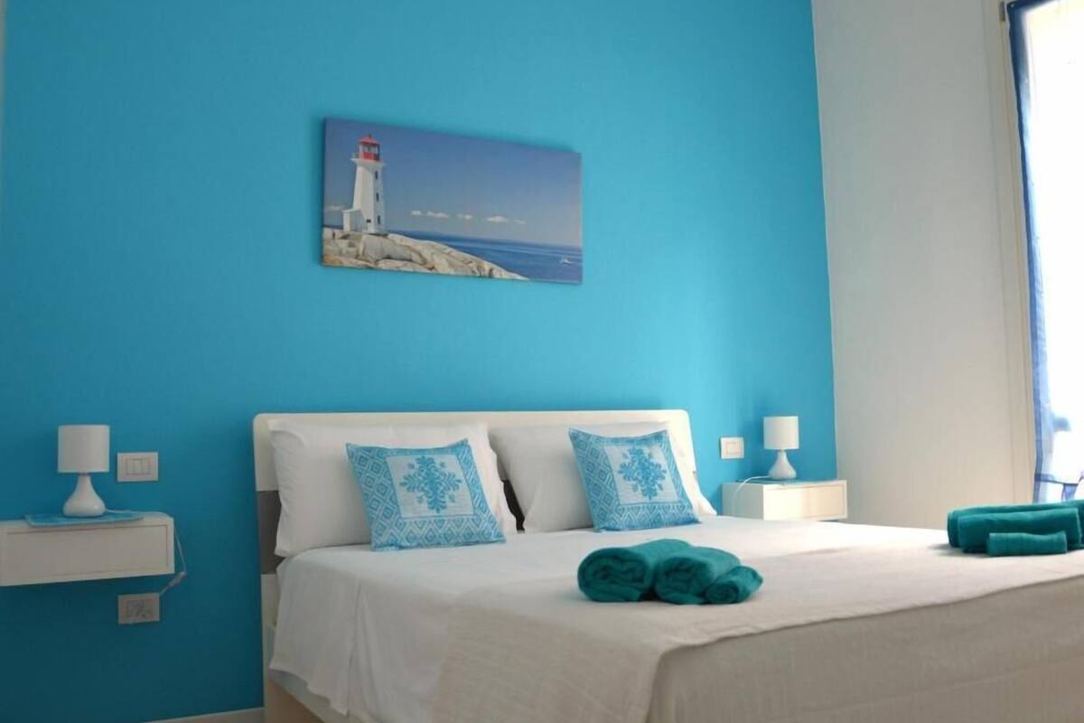 Quarto com cama, almofadas, pintura de um farol e janela.