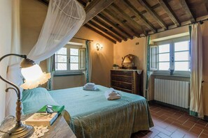 Ein Schlafzimmer mit Bett, Nachttisch, Nachttischlampe, Kommode, Spiegel und einem Fenster mit Vorhängen.