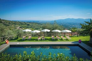 Ein Pool mit Liegestühlen und Sonnenschirmen, umgeben von Grün und Blick auf ferne Hügel.