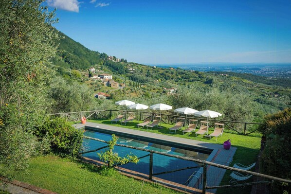Zona de piscina con sillas de descanso y sombrillas, rodeada de vegetación y con vistas a un valle.