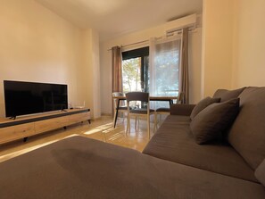 Ein Wohnzimmer mit Flachbildfernseher auf einem Holz-TV-Schrank, einer Couch, einem Esstisch mit Stühlen und einem Fenster mit Vorhängen.