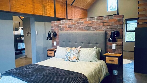 Een slaapkamer met een bakstenen muur, een bed met een grijs hoofdeinde, nachtkastjes en een raam.