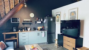 Een compacte keuken met een magnetron, koffiezetapparaat en koelkast. Er staat een houten tafel met een boek en een fles, en een klein krukje.