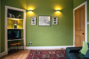 Een woonkamer met een televisie, een boekenkast, ingelijste foto's en een kamerkleed met een patroon.