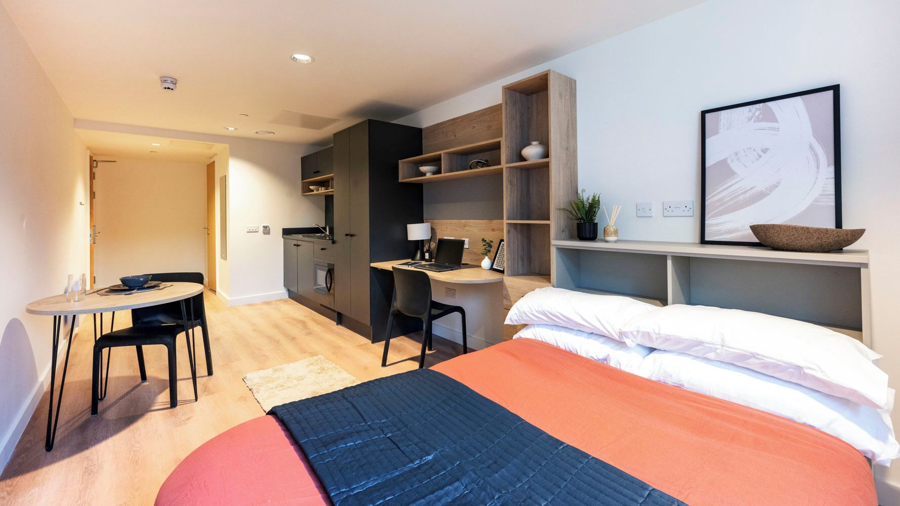 A modern hotel room with a bed, desk, chair, kitchenette, and a small dining table.