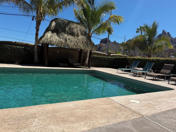 A swimming pool with lounge chairs, a thatched-roof structure, and palm trees.