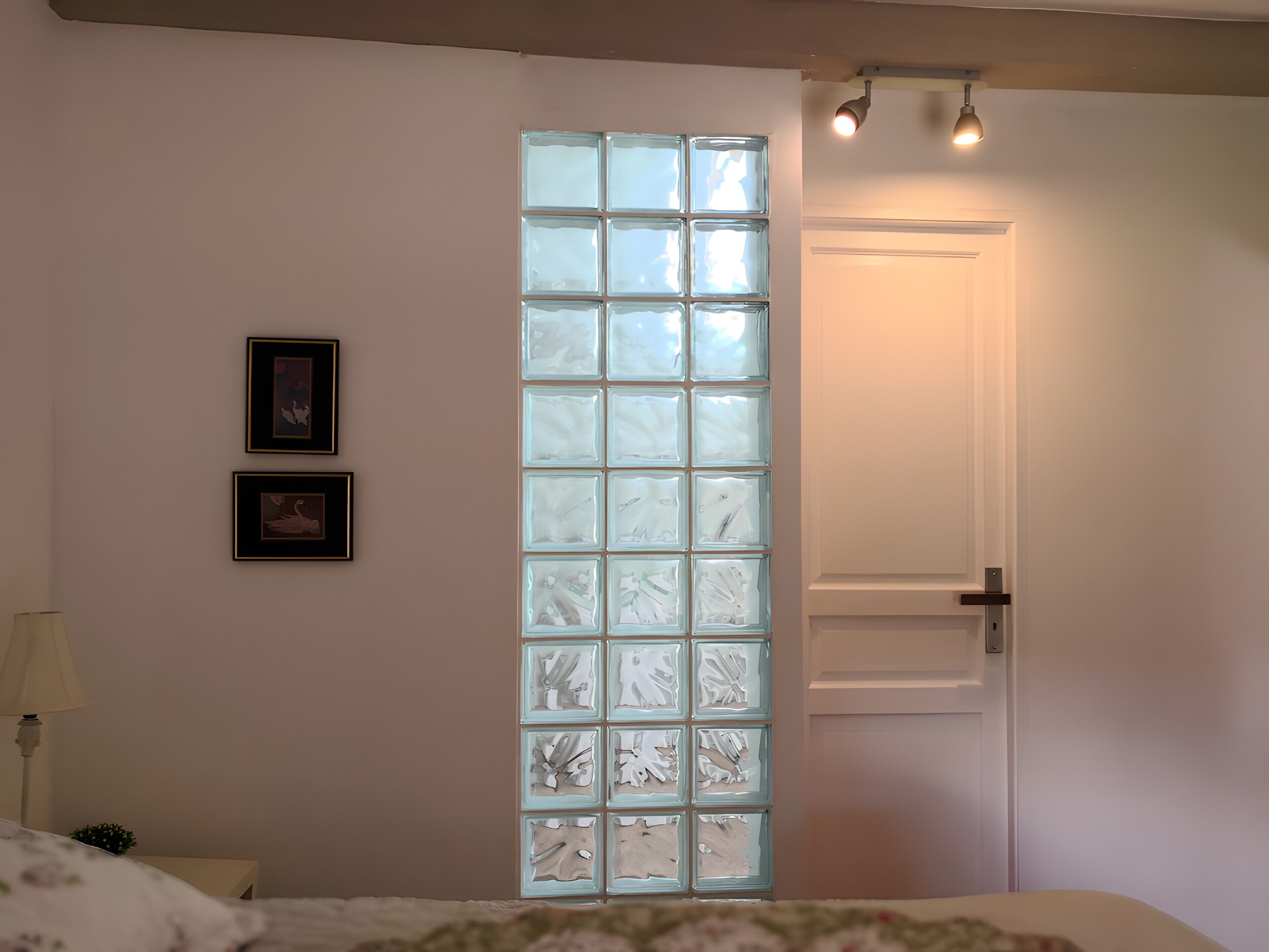 Ein Schlafzimmer mit einem Bett, einem Nachttisch, einer Lampe, einem an der Wand befestigten Regal mit Bildern und einer Tür mit Milchglasfenster.
