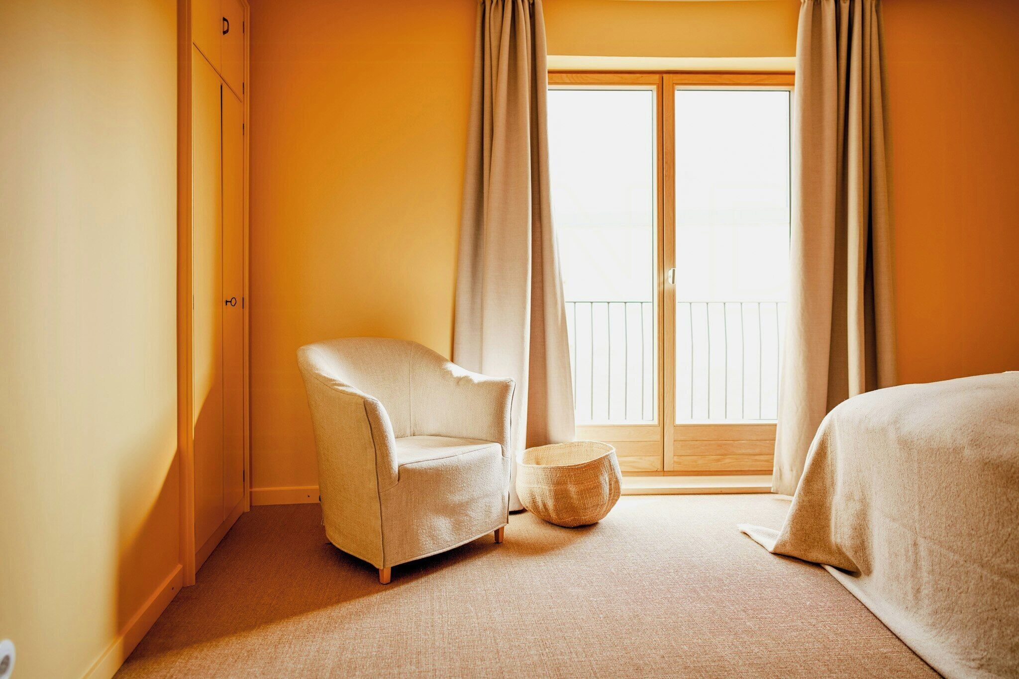 Une chambre d’hôtel avec un lit simple, une chaise, un pouf rond et une porte-fenêtre coulissante donnant sur l’extérieur.