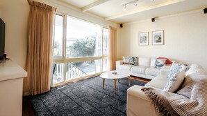 Une pièce avec un canapé blanc, une petite table ronde et un téléviseur posé sur un meuble. De grandes fenêtres sont présentes, ainsi que des rideaux, un balcon et deux tableaux encadrés accrochés au mur.