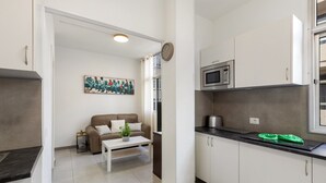 Cozinha moderna com armários brancos, micro-ondas e máquina de café. Há um sofá marrom e uma pequena mesa com uma planta.