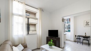 Uma sala de estar com sofá, uma mesa de centro com uma planta, televisão e um espaço de jantar com cadeiras e mesa.