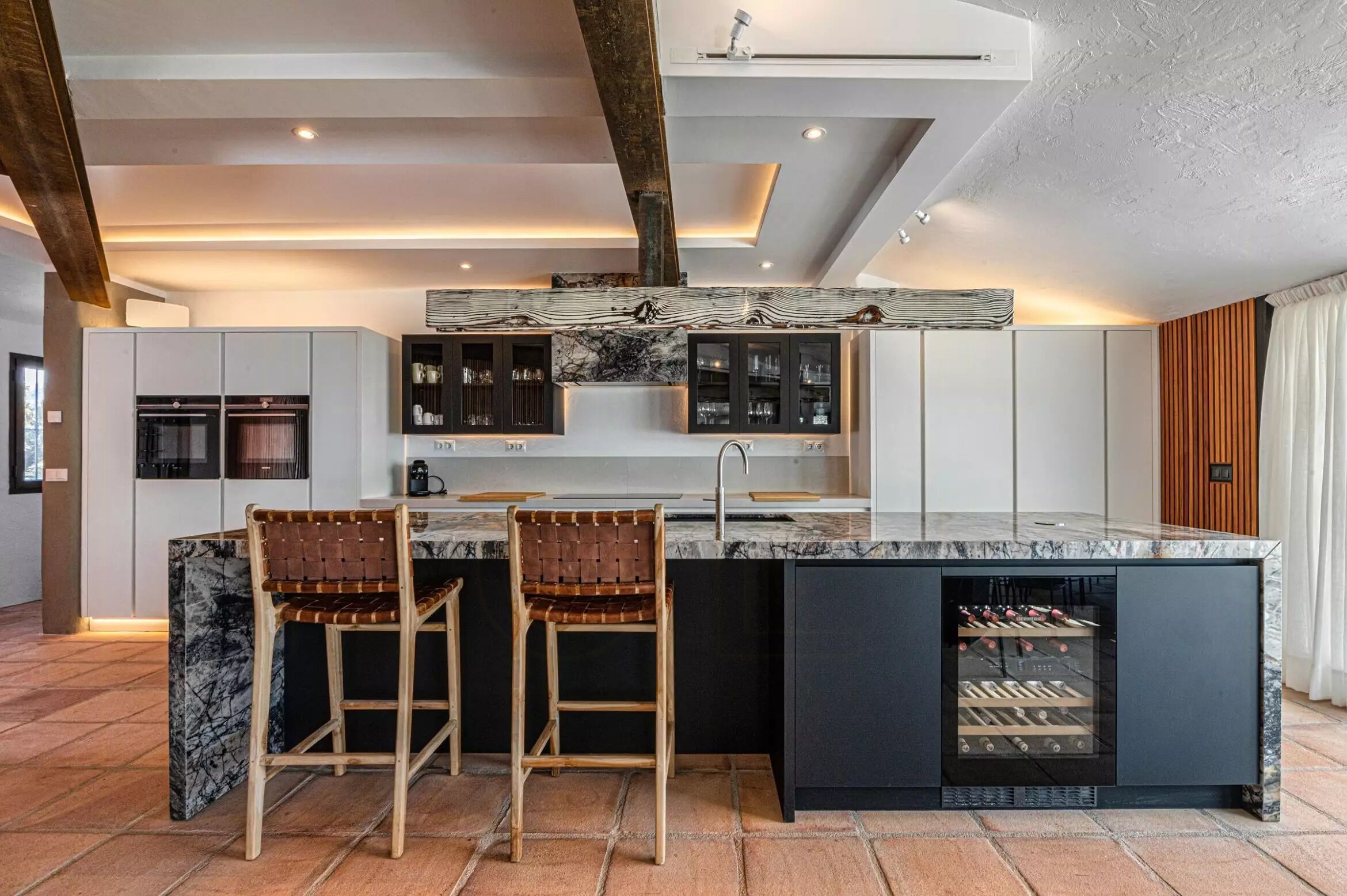 Een moderne keuken met een centraal eiland, barstoelen, een ingebouwde wijnkoeler en een groot raam.